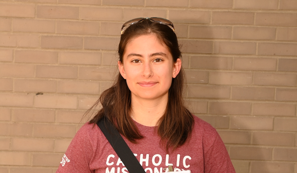 A woman with shoulder-length brown hair, wearing a Catholic University sweatshirt, stands in front of a brick wall.