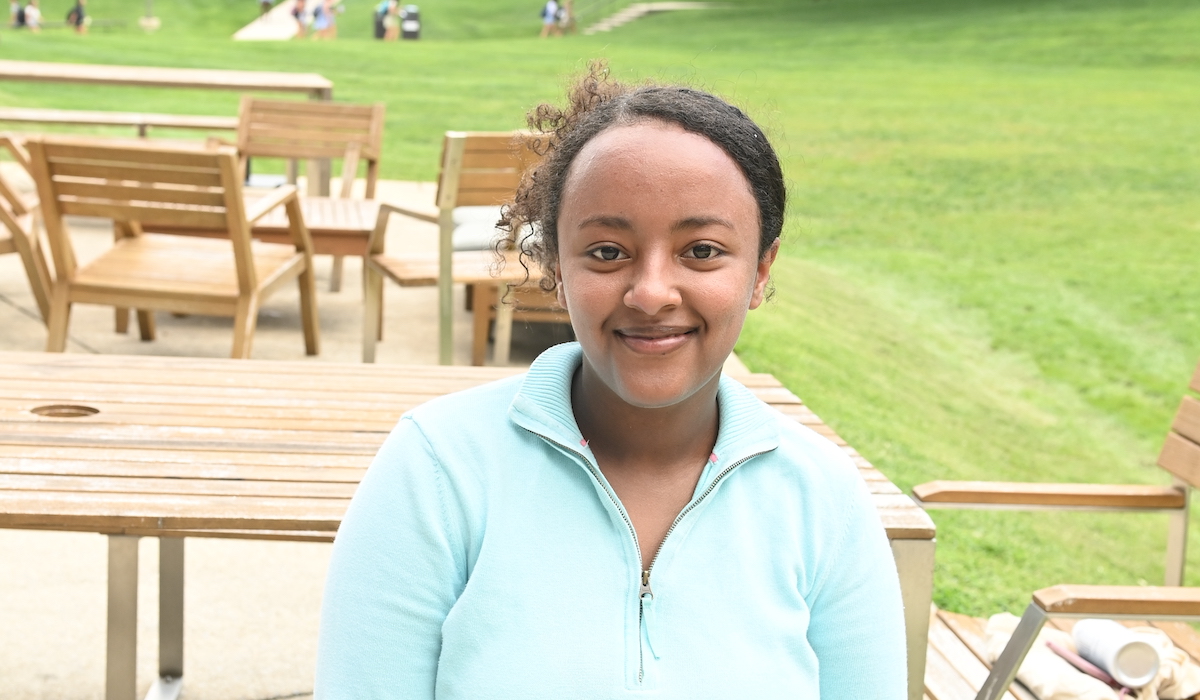 A woman is standing in front of a sprawling lawn darted with wooden chairs. Her curly hair is up in a bun and she is wearing a mint 3/4 zip sweater.