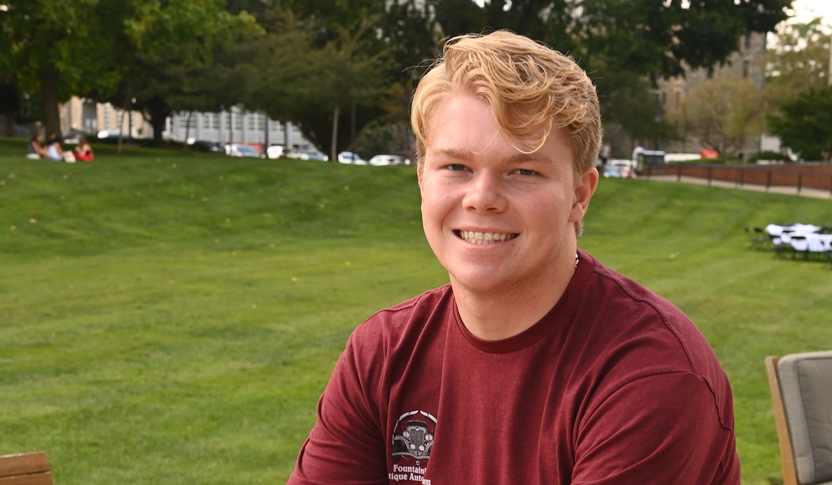 A man with blonde hair and a wide smile, wearing a red shirt, sits in front of a lush green lawn and trees.