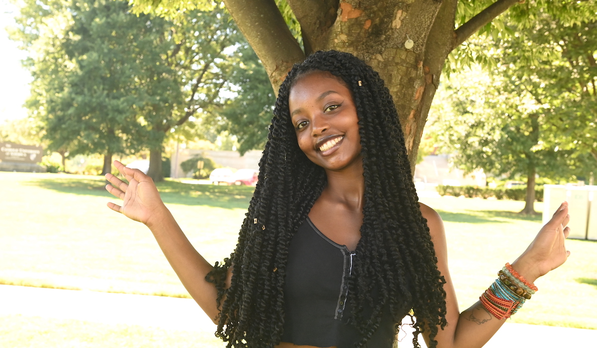 A woman in a black tank top and outstretched arms is standing in front of a tree