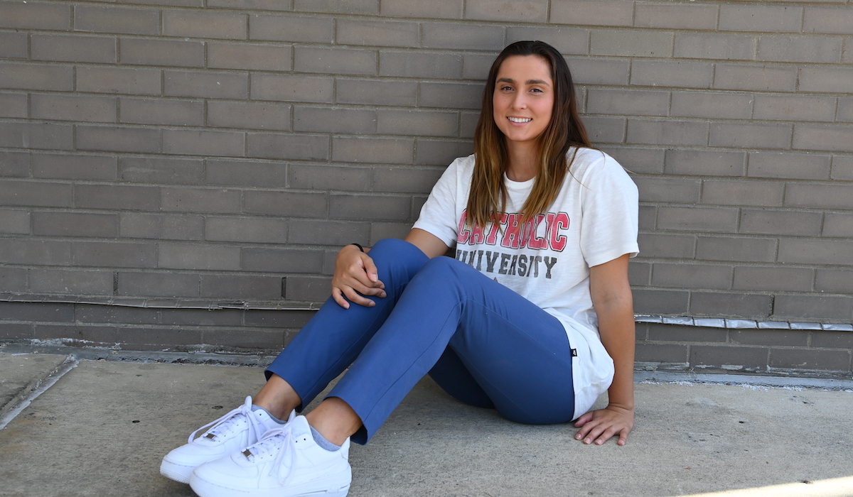 A woman sits on the ground and leans against a brick wall. She is wearing a Catholic University tee shirt, jeans, and white sneakers. She has long brown hair.