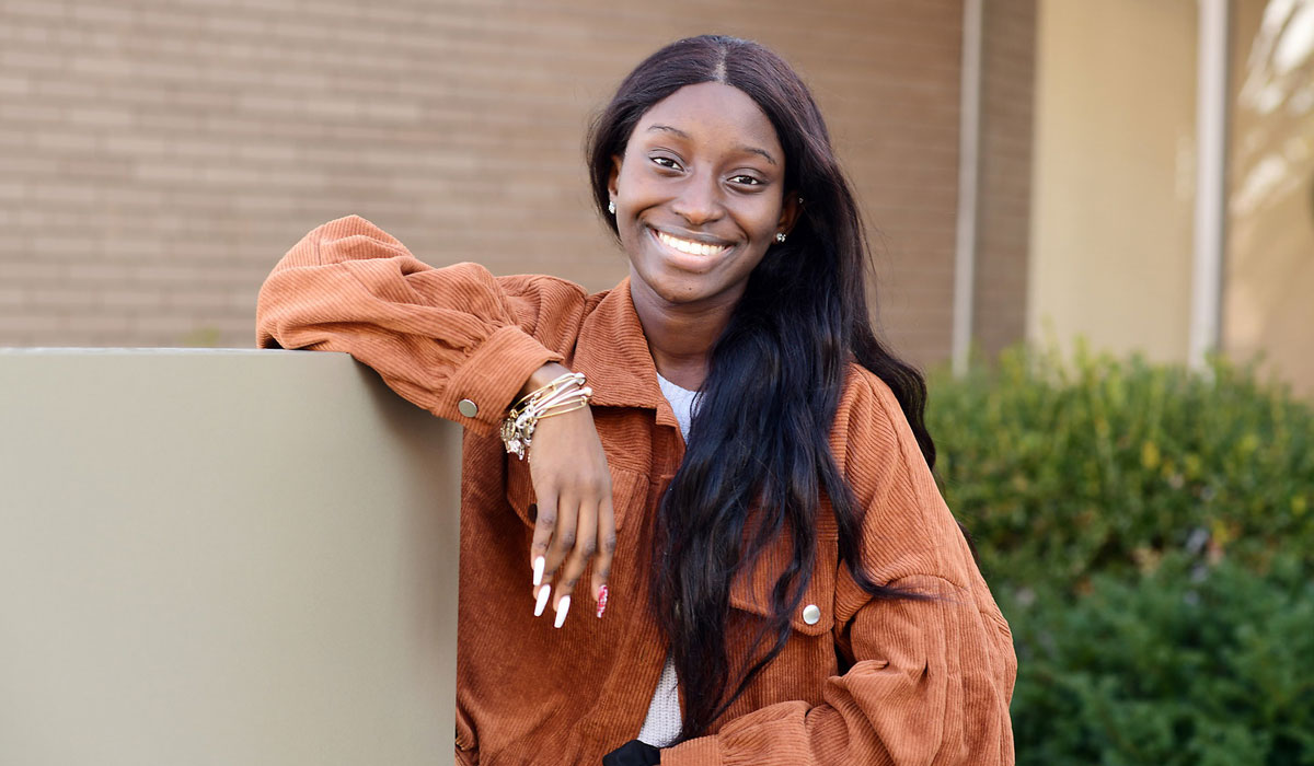 A woman is standing in front of a brick wall and hedges. She is leaning against a shorter all. She has long black hair and is wearing an orange corduroy jacket over a white tee-shirt. She has silver bracelets on her propped wrist.