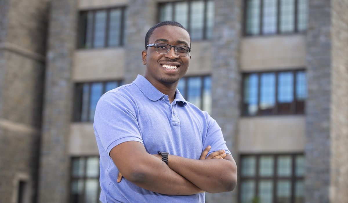 A man stands in front of a grey brick building. He is wearing a blue shirt and has glasses.