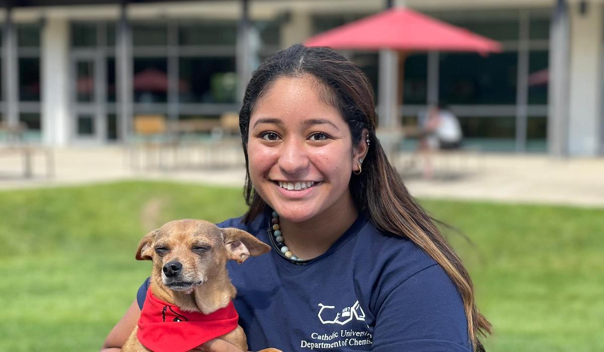 A woman with long brown hair and a blue shirt stands in front of the Pryz Center at Catholic University. She's holding a small dog wearing a red bandana.