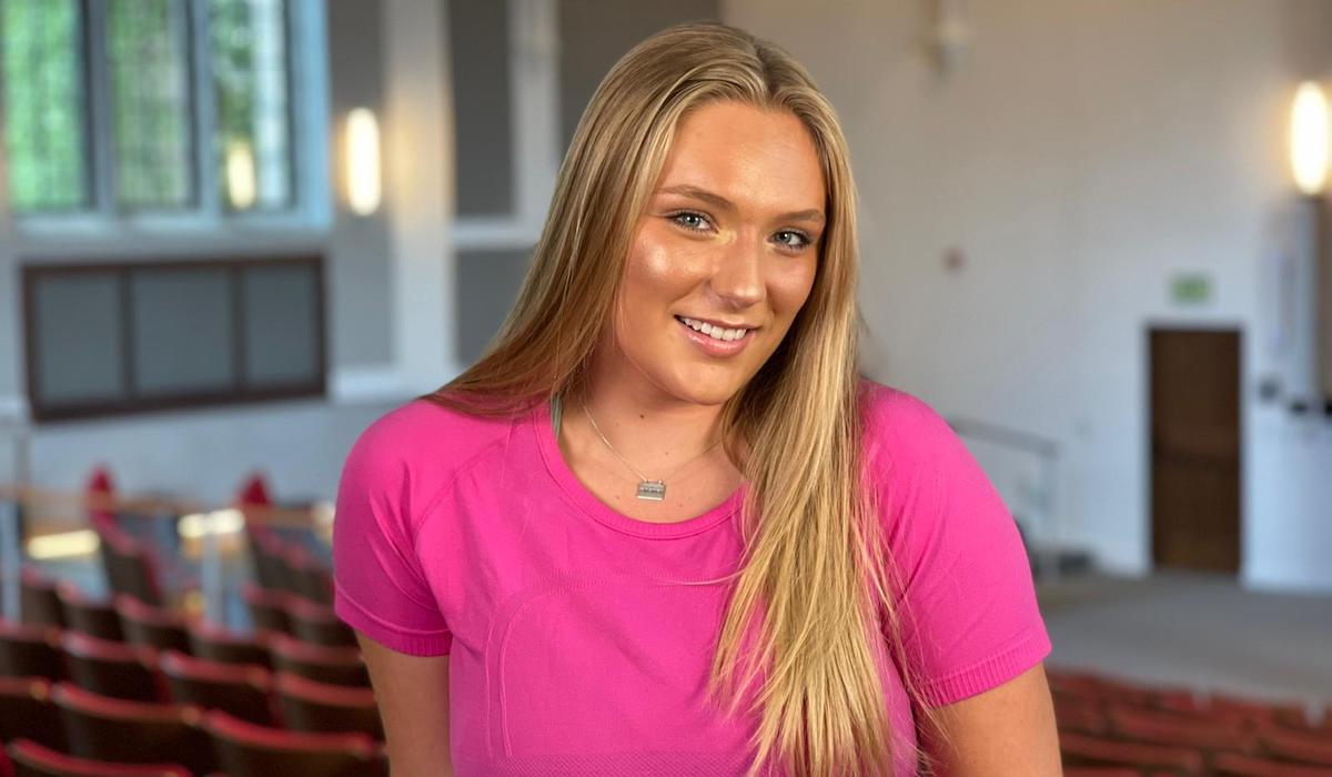 A woman stands in the back of an auditorium. She is wearing a bright pink shirt and has long blonde hair. 