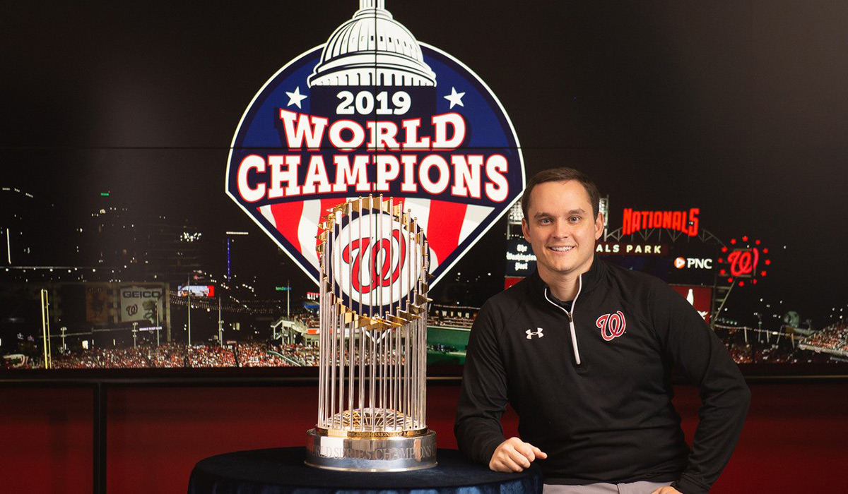 A man in a Washington Nationals 3/4 zip sweatshirt is standing in front of Nationals Park at night. There is a sign that says "2019 World Champions" directly behind him. 