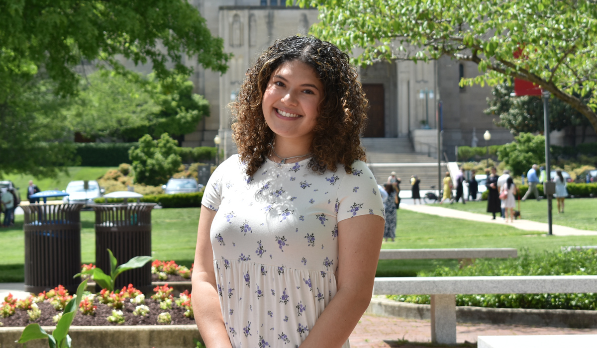 A woman with curly brown hair stands in front of a courtyard featuring a lush lawn, benches, trees, and the entrance to a cathedral. She is wearing a floral top.