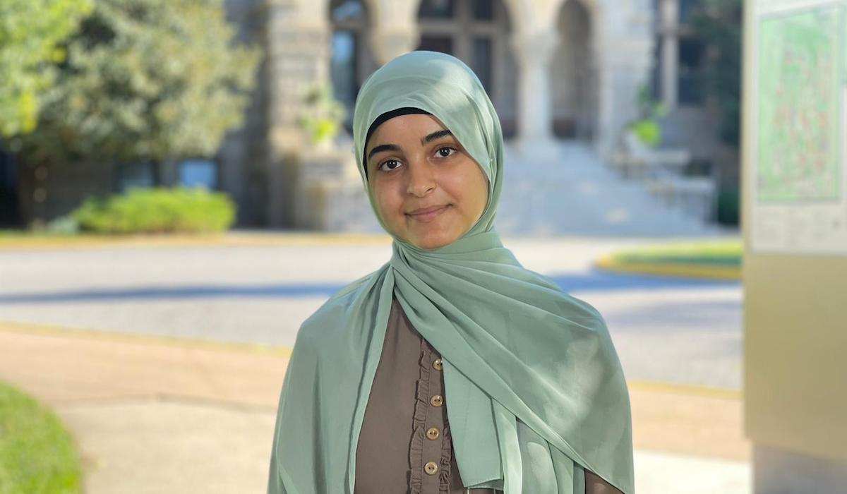 A woman stands in front of a court yard with a stone building and trees. She is wearing a grey dress and a mint green hijab