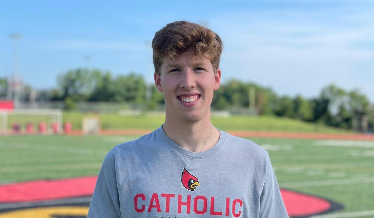 A man with curly blonde hair in a Catholic University shirt stands in front of a football field.