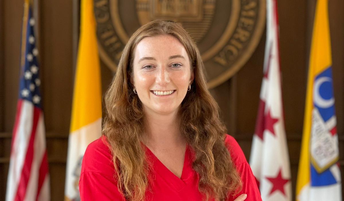 A woman with long red-blonde hair stands in front of The Catholic University of America's crest in Heritage Hall. Behind her are four flags, including the flag of the United States, the Vatican, and Washington, D.C.