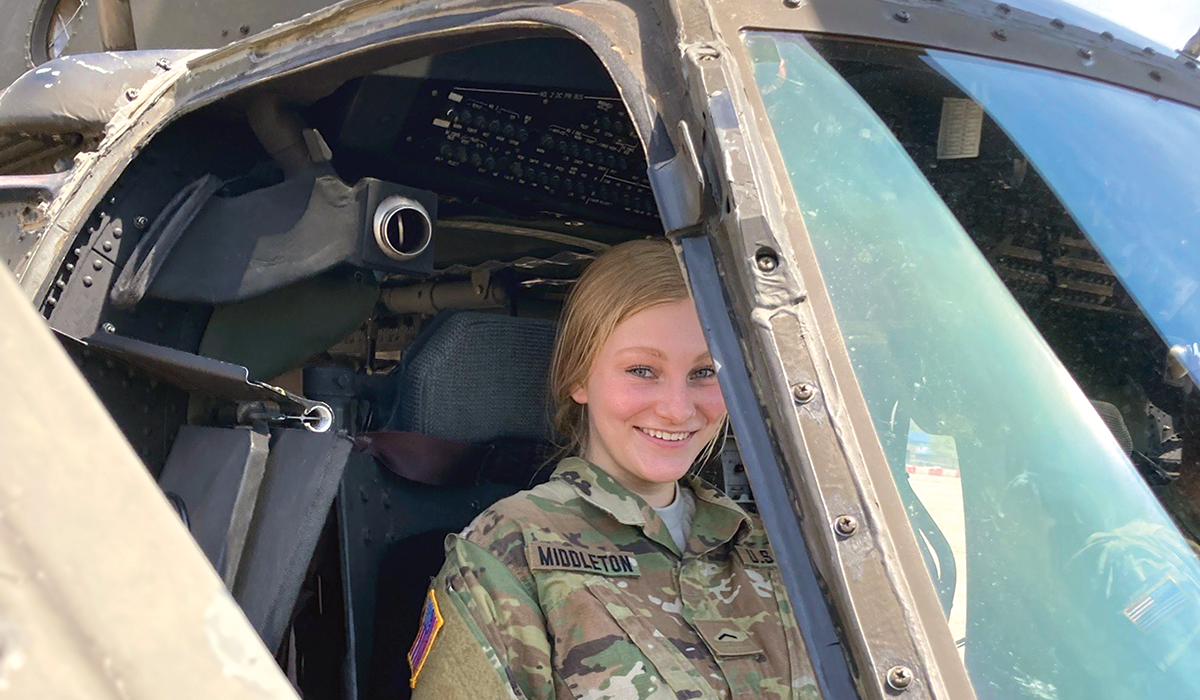 A woman is in the cockpit of a helicopter. She had blonde hair that is tucked into a bun and is wearing army fatigues with her last name, MIDDLETON, stitched on the pocket. 