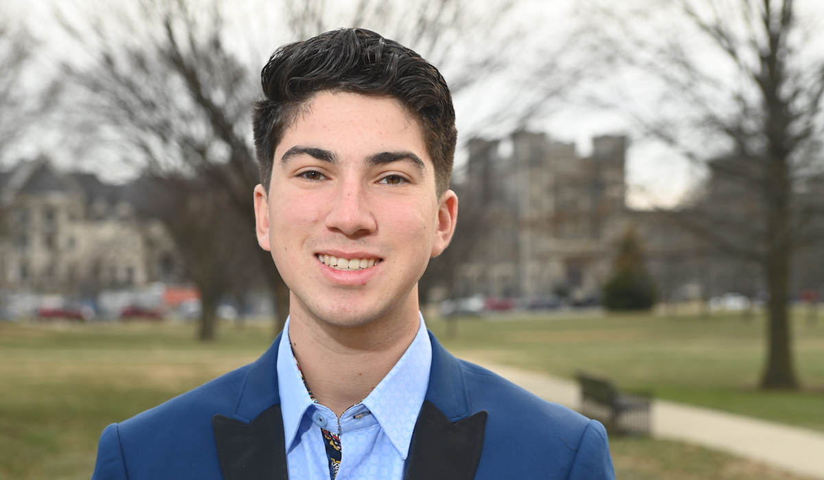 A man stands in front of Catholic University's campus. He is wearing a blue suit jacket over a light blue button down shirt.