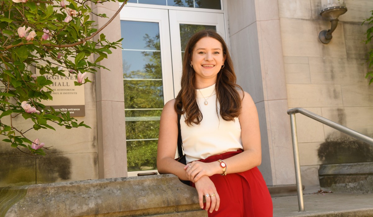 Julia Englert is standing at the Busch School of Business, leaning against a podium. She has long brown hair and is wearing a white top and red pants. 