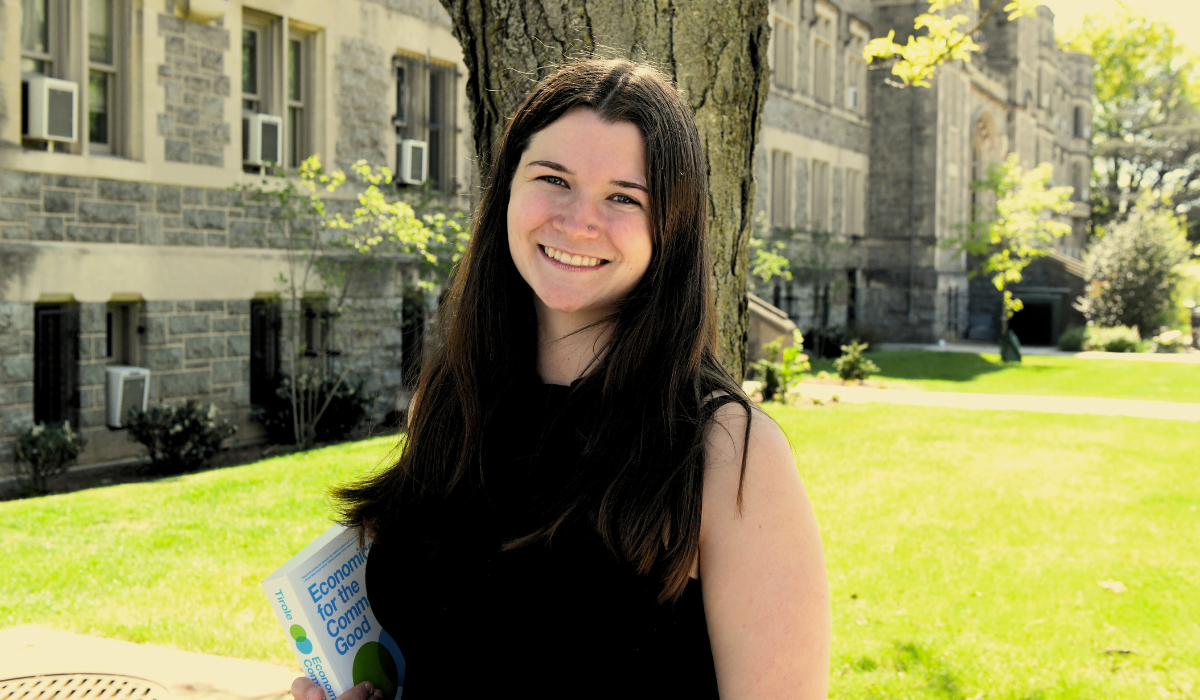 Maria Duffy, stands in front of O'Connell Hall in spring time. She has dark hair that reaches past her shoulders and is wearing a black sleeveless top. She is holding a book.