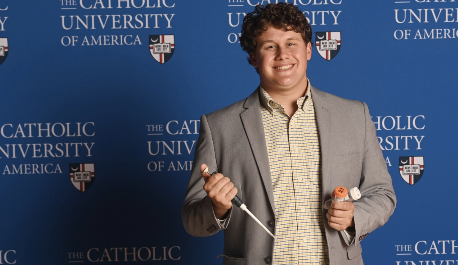 Elijah is standing in front of a Catholic University background. He has curly hair and is wearing a checkered shirt and a grey blazer.