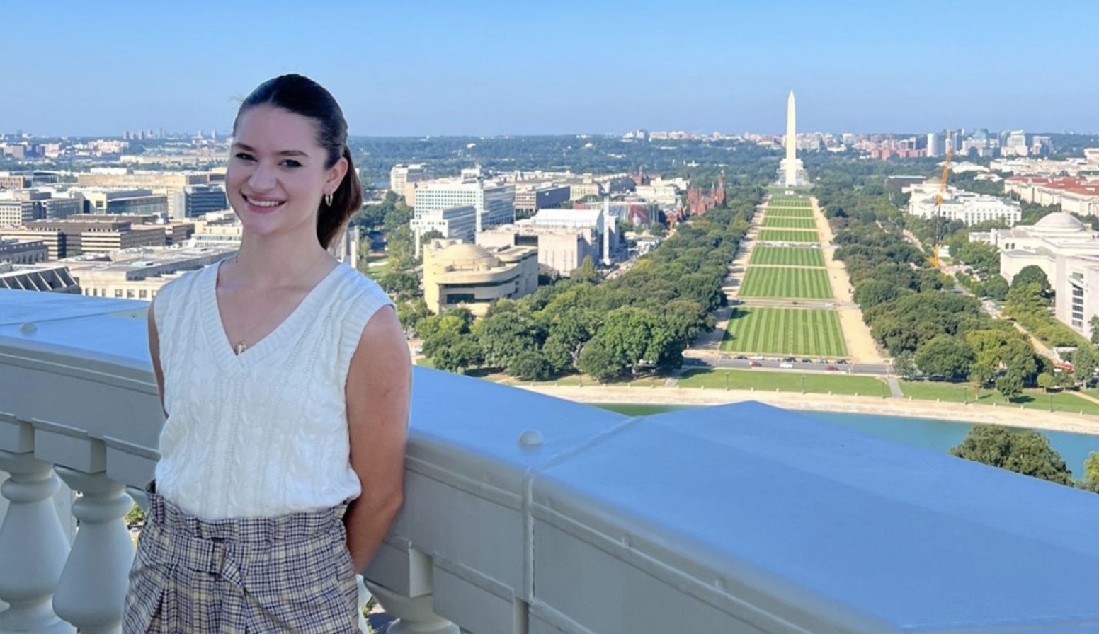 Julia Englert is standing at the Capitol's balcony, looking over the National Mall. She is wearing a white shirt and plaid pants. 