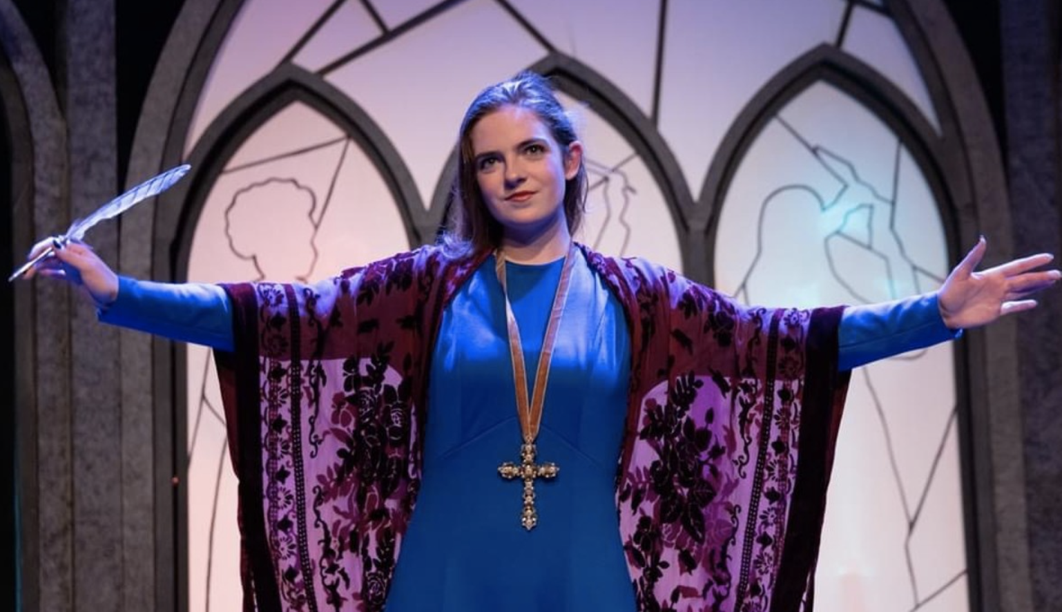 Kathryn Parr is performing in a stage production. She is standing against illuminated windows, her arms wide and draped in a red lace shawl. She is also wearing a blue dress and a large cross. In her right hand, she has a feather pen.