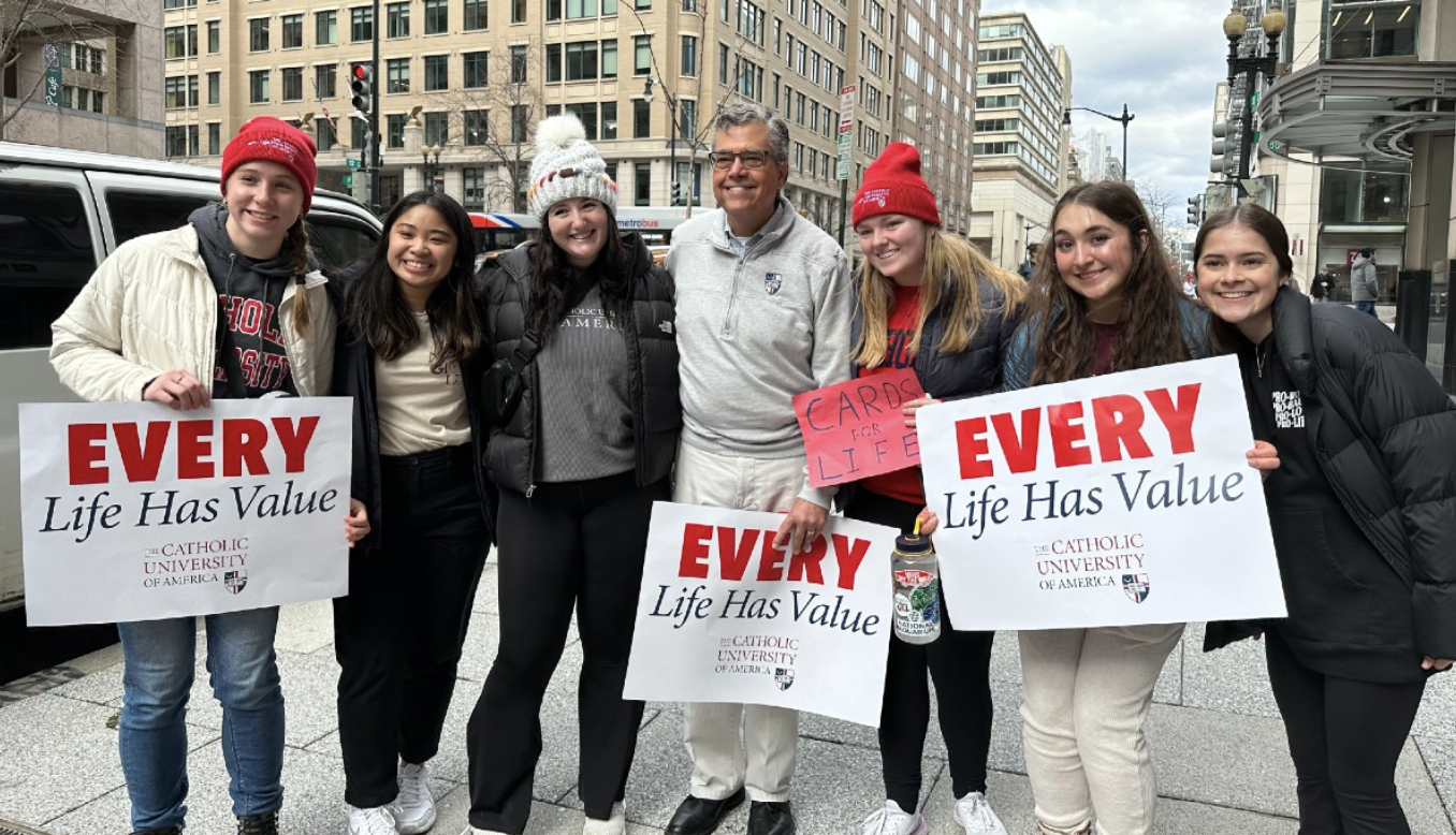 A group of six female Catholic University Students stand with President Peter Kilpatrick downtown. They are all dressed for the winter and are holding "Every Live Has Value" signs.