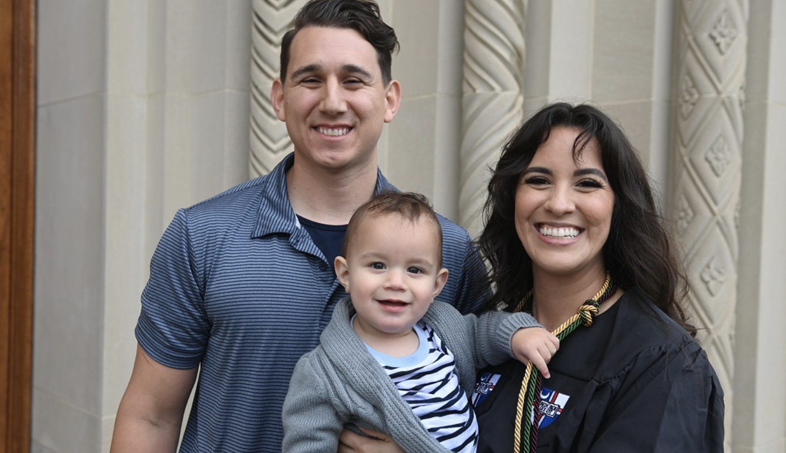 Cassandra is standing with her family in front of a building entrance. She is wearing CUA graduation regalia while holding a baby. A man wearing a blue polo shirt is standing next to her.