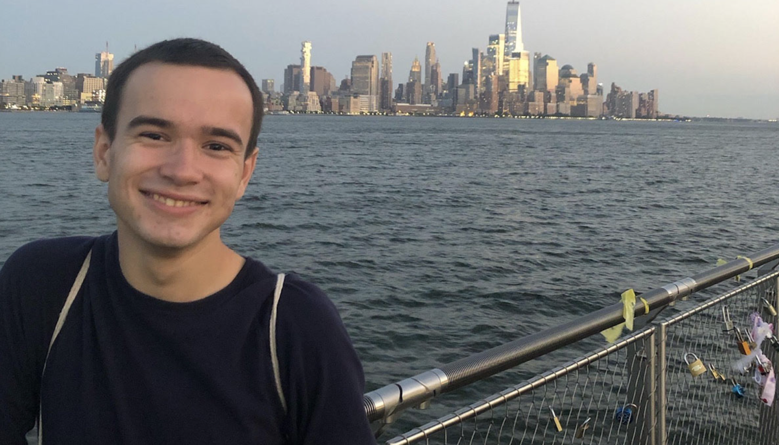 Benjamin Duphiney stands in front of the New York City skyline. He is wearing a blue sweater and has short dark hair.