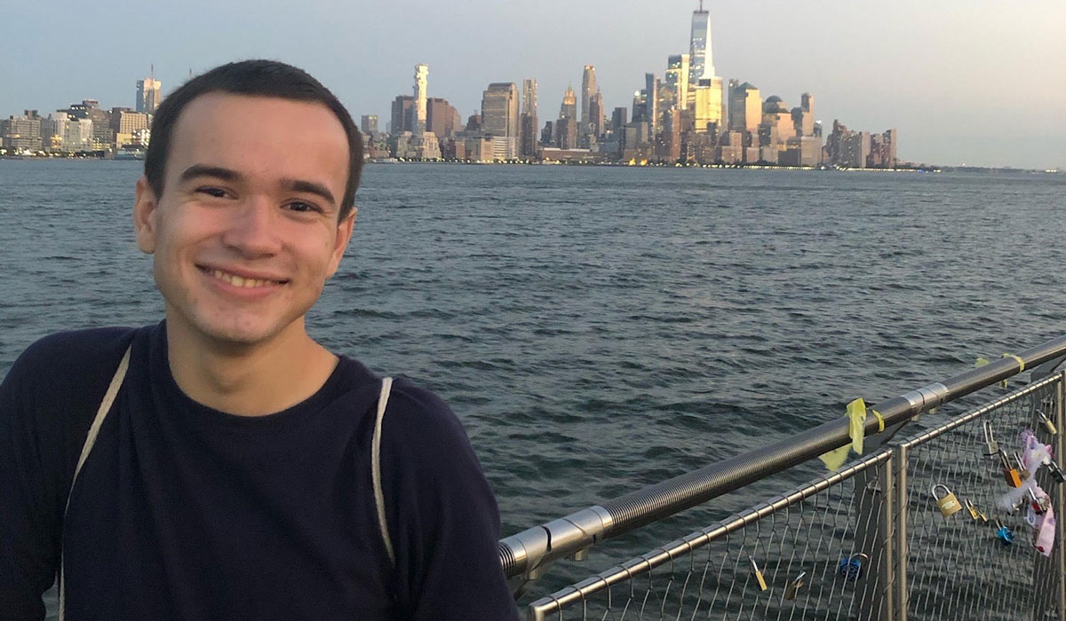 A man is standing on a bridge that overlooks the New York City skyline and harbor. He is wearing a plain navy blue sweatshirt and has very short dark hair.