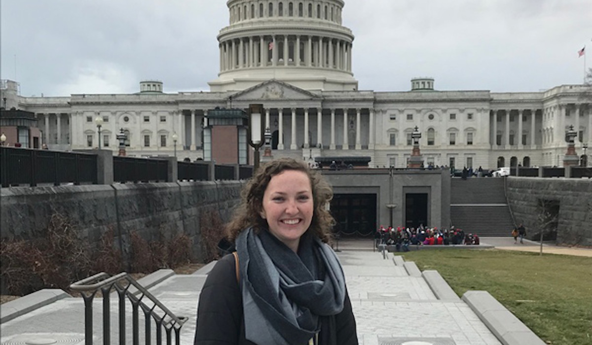 A woman is standing in front of the US Capitol. She has a long curly hair and is wearing a dark grey shirt and a large scarf.