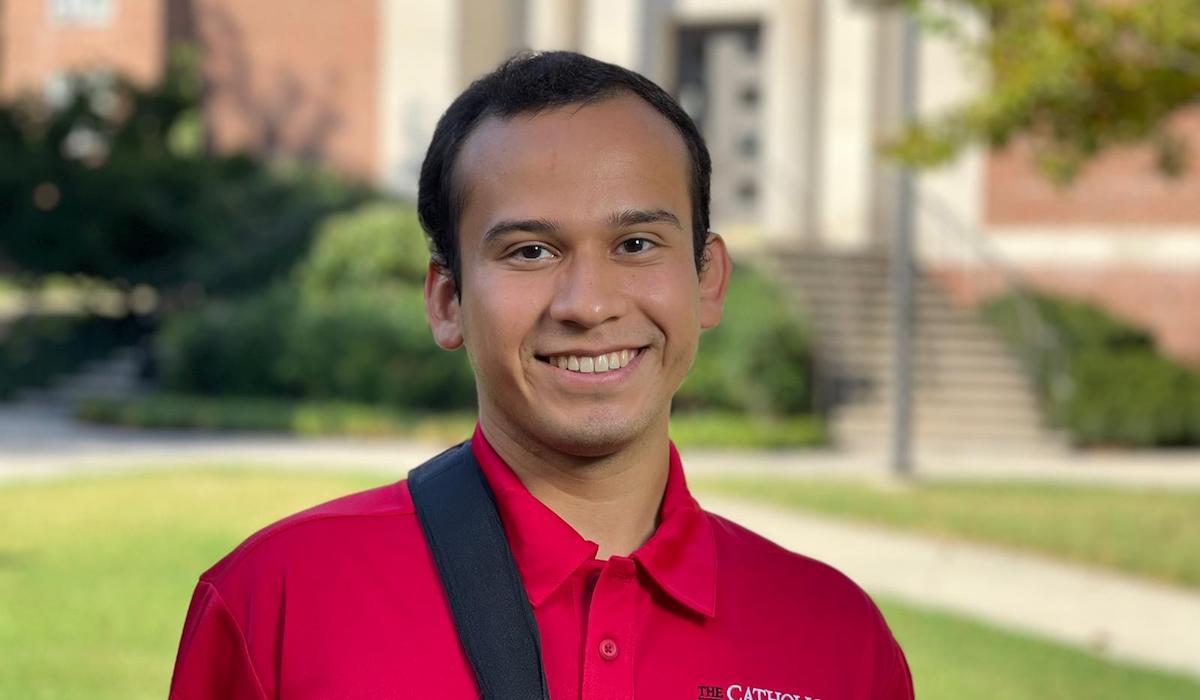 A man is wearing a red polo in front of a red brick building, a bush, and a lush lawn. 