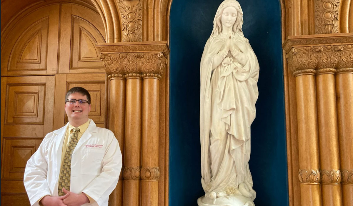 A man is standing in a church next to a Virgin Mary statue. He is wearing a white coat, a light yellow shirt, and a dark yellow tie. 