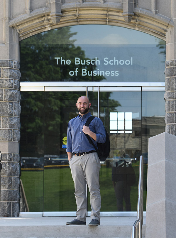 A man with a beard is standing in front of The Busch School of Business' glass doors. He is wearing khaki pants, a deep blue shirt, and has a backpack slung over his shoulder.