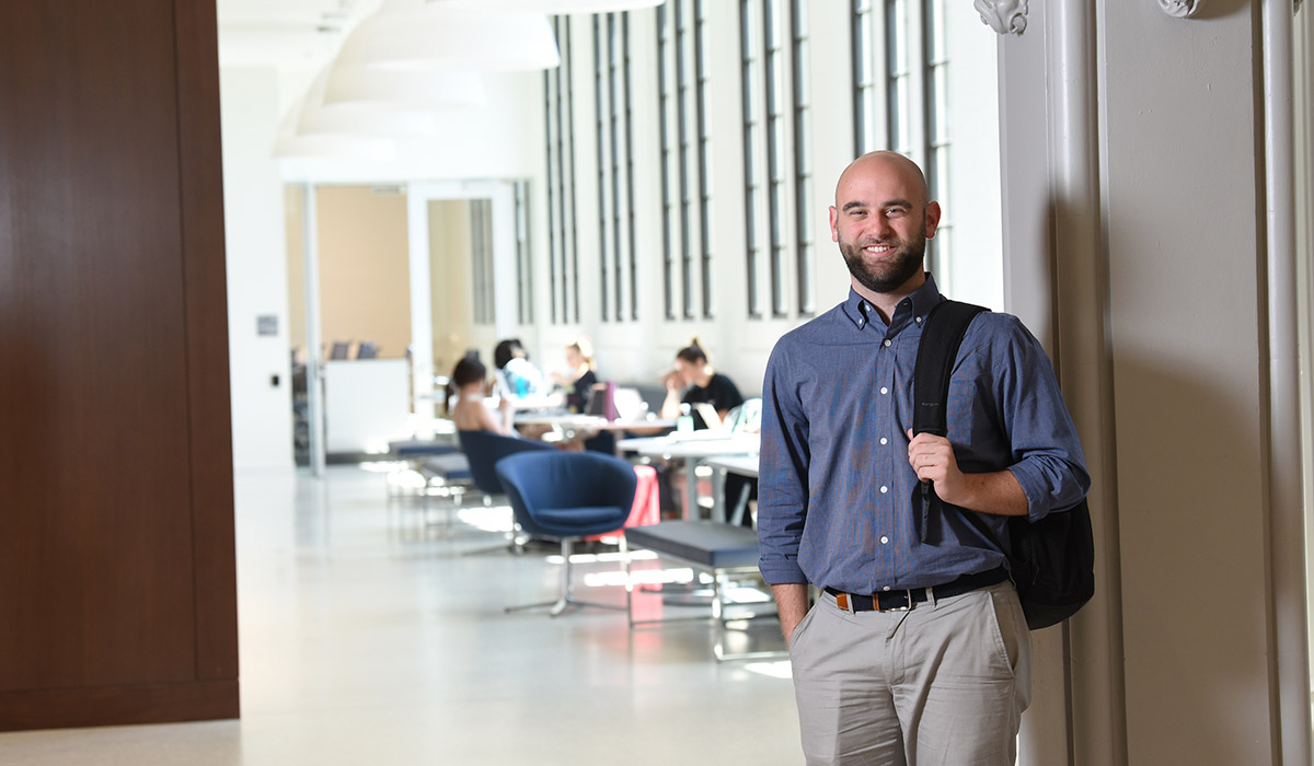 A man is standing in a classroom. Behind him are several students studying in a group. He is wearing a blue shirt and khaki pants. A black backpack is slung over his shoulder.