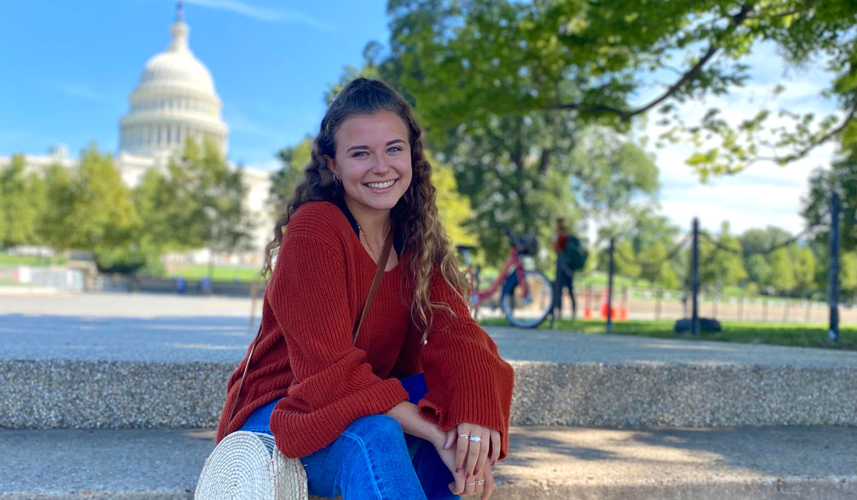 A woman is sitting in front of the US Capitol Building. She is wearing jeans and a long-sleeve orange blouse. She has long curly brown hair.