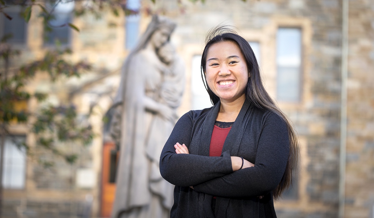 A woman with long black hair is standing with her arms crossed in front of a statue of Mary and Jesus and grey brick building. She is wearing a black cardigan and a red shirt.