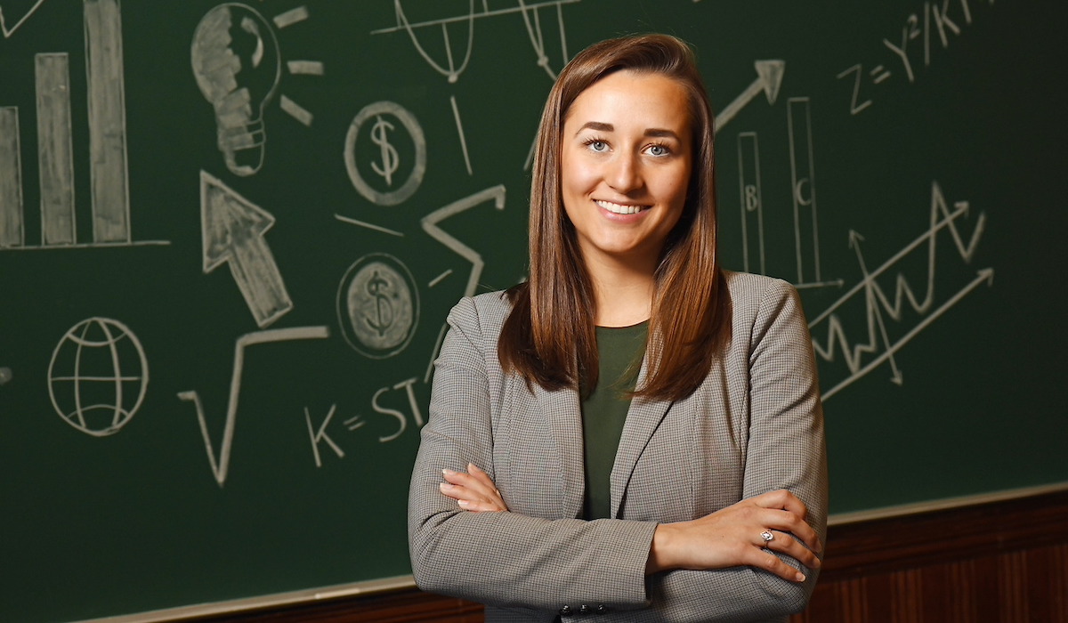 A woman is standing with her arms crossed in front of a blackboard covered in economic graphs. She has shoulder length brown hair and is wearing a grey blazer over an olive green shirt. 