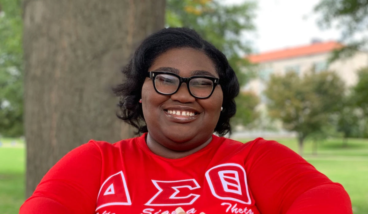 A woman is standing in front of a tree and a building. She has short dark hair and is wearing a red shirt and glasses.