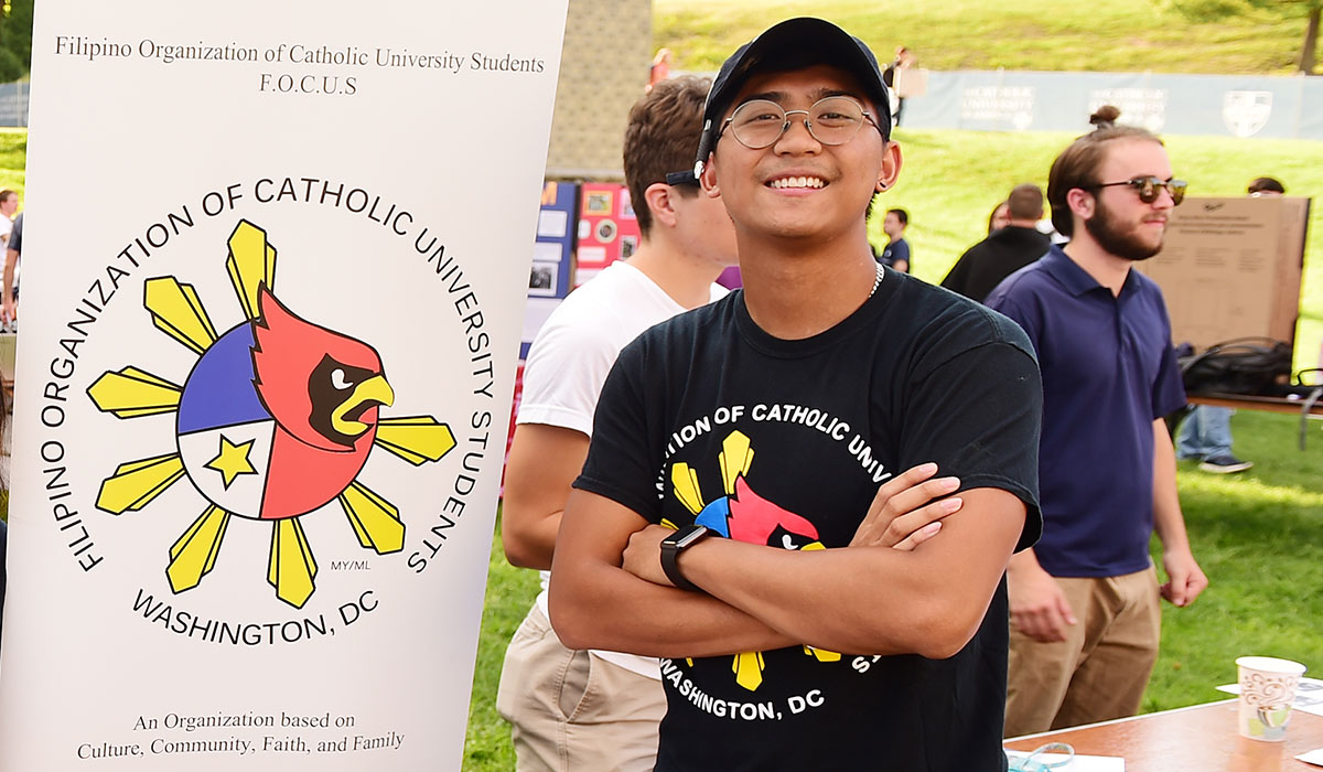 A man is standing with his arms crossed at an activities event on CUA Campus. He is wearing a Filipino Organization of Catholic University Students (FOCUS) which features a Cardinal logo and the flag of the Philippines. Behind him is a sign with the same design.