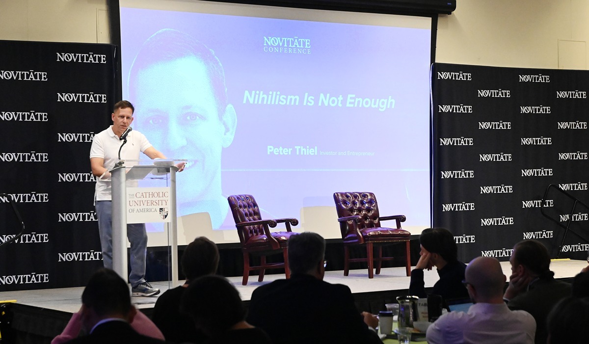 Peter thiel on stage speaking at a podium in front of a screen with the title "nihilism is not enough"