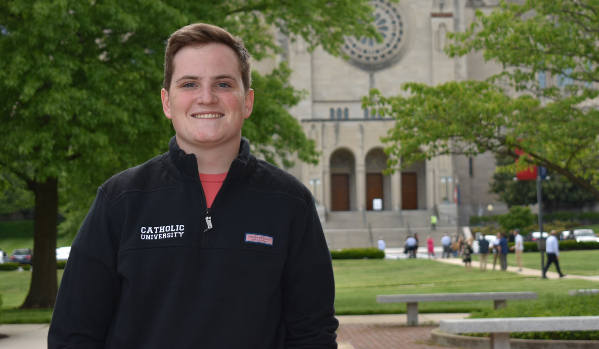 A man wearing a Catholic University 3/4 zip sweatshirt is standing in front of The Basilica of the National Shrine of the Immaculate Conception.