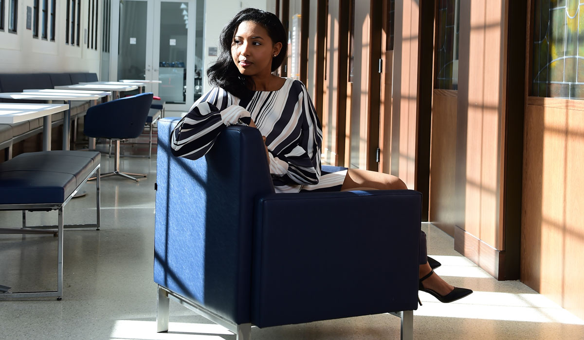 A woman in a black and white dress is sitting over the back of a navy blue chair. The chair is in the middle of a hallway in The Busch School of Business.