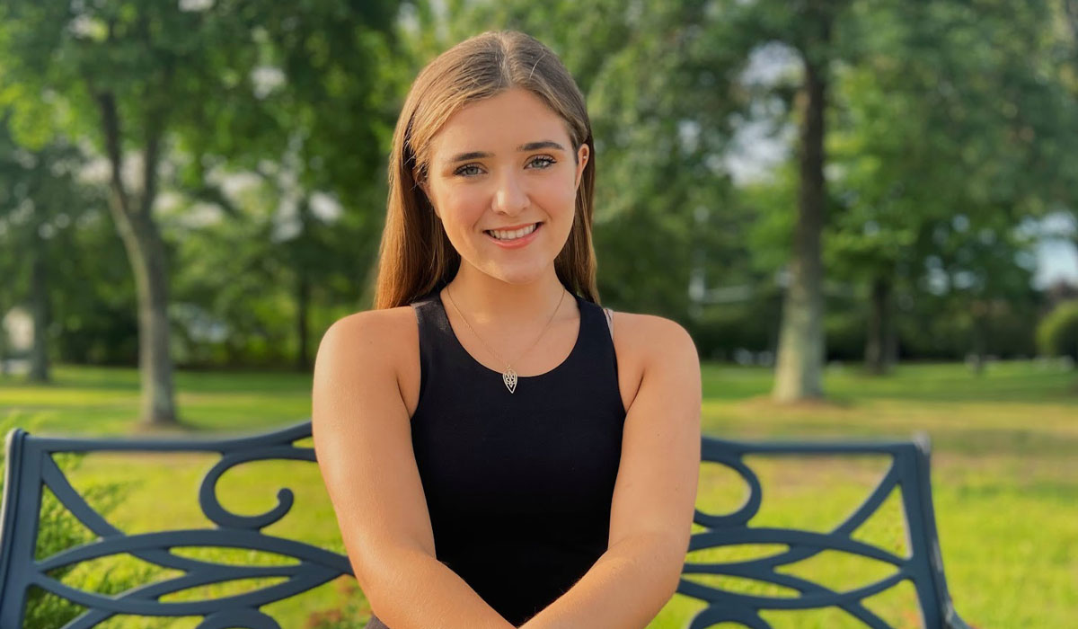 A woman is sitting on a bench in front of a lush lawn and trees. She has long blonde hair and is wearing a black tank top. She has a silver necklace around her neck.