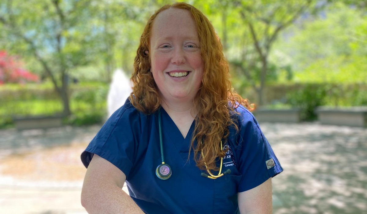 Mairin stands in front of green courtyard. She has long curly red hair and blue scrubs labeled "The Catholic University of America." A stethoscope is slung over her shoulder. 