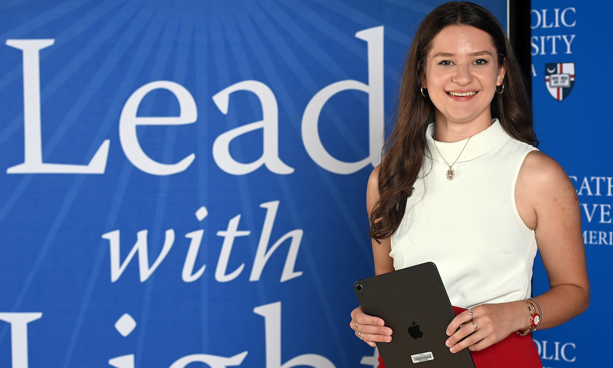 Julia Englert is standing in front of a Catholic University Lead With Light background. She has long brown hair and is wearing a white top and red pants. 