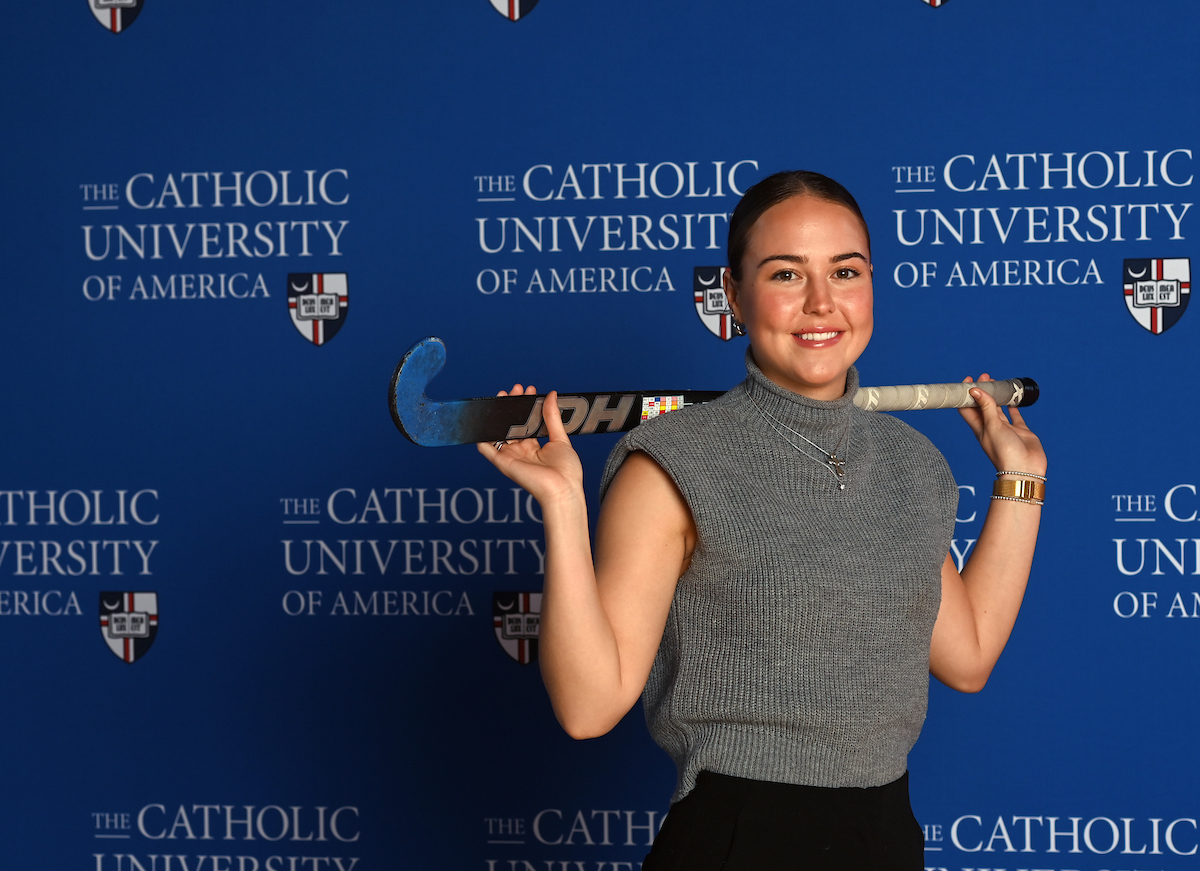 Mary Parker is standing in front of a blue Catholic University of America background. She has her field hockey stick draped over her shoulder.