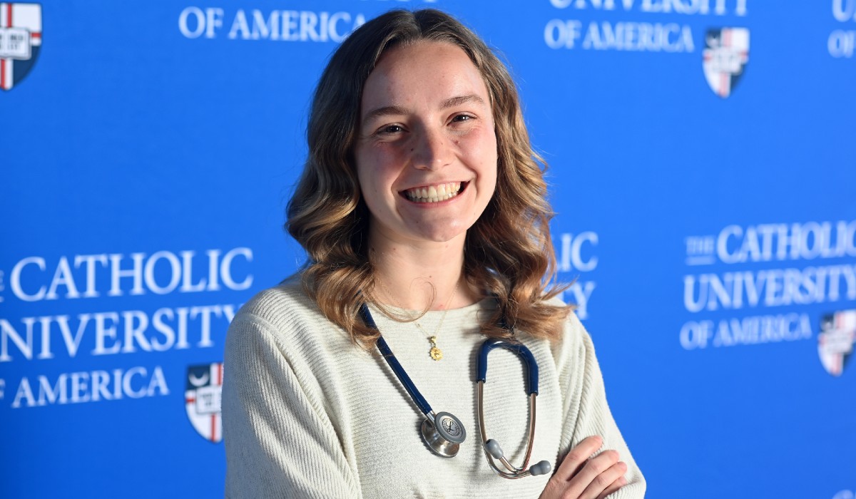 Elizabeth is standing in front of a blue Catholic University background. She is wearing white and has a stethoscope around her neck.