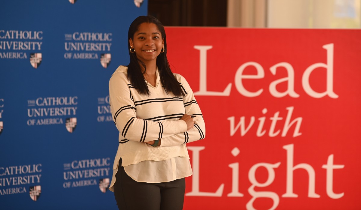 Lauren White is standing in front of blue and red Catholic University backdrops. Her arms are crossed and she's smiling. She is wearing a white and black sweater and dark pants.