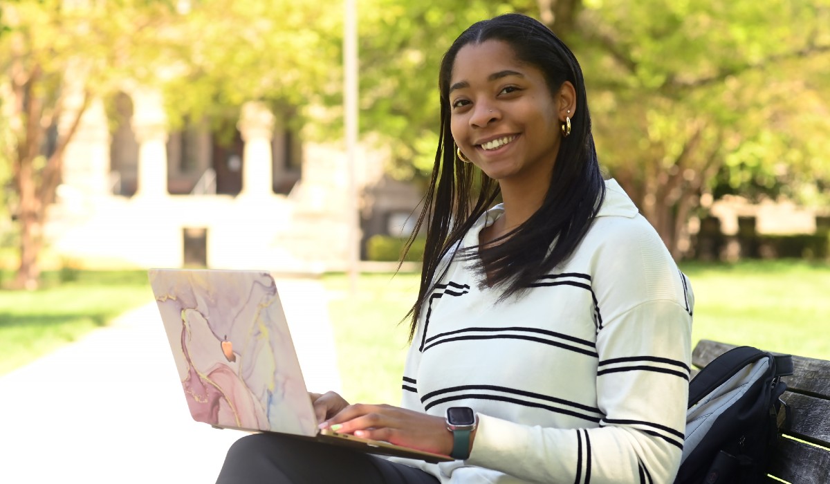 Lauren White is sitting on a bench on Catholic University's campus, with a laptop perched on her knees. She is wearing a white and black sweater and dark pants.