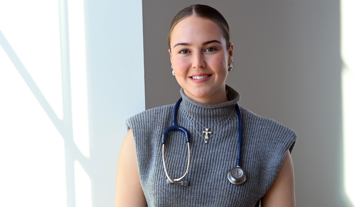 Mary is standing in front of a window in the Conway School of Nursing. She is wearing a grey sweater. Around her neck is a silver cross and a stethoscope.