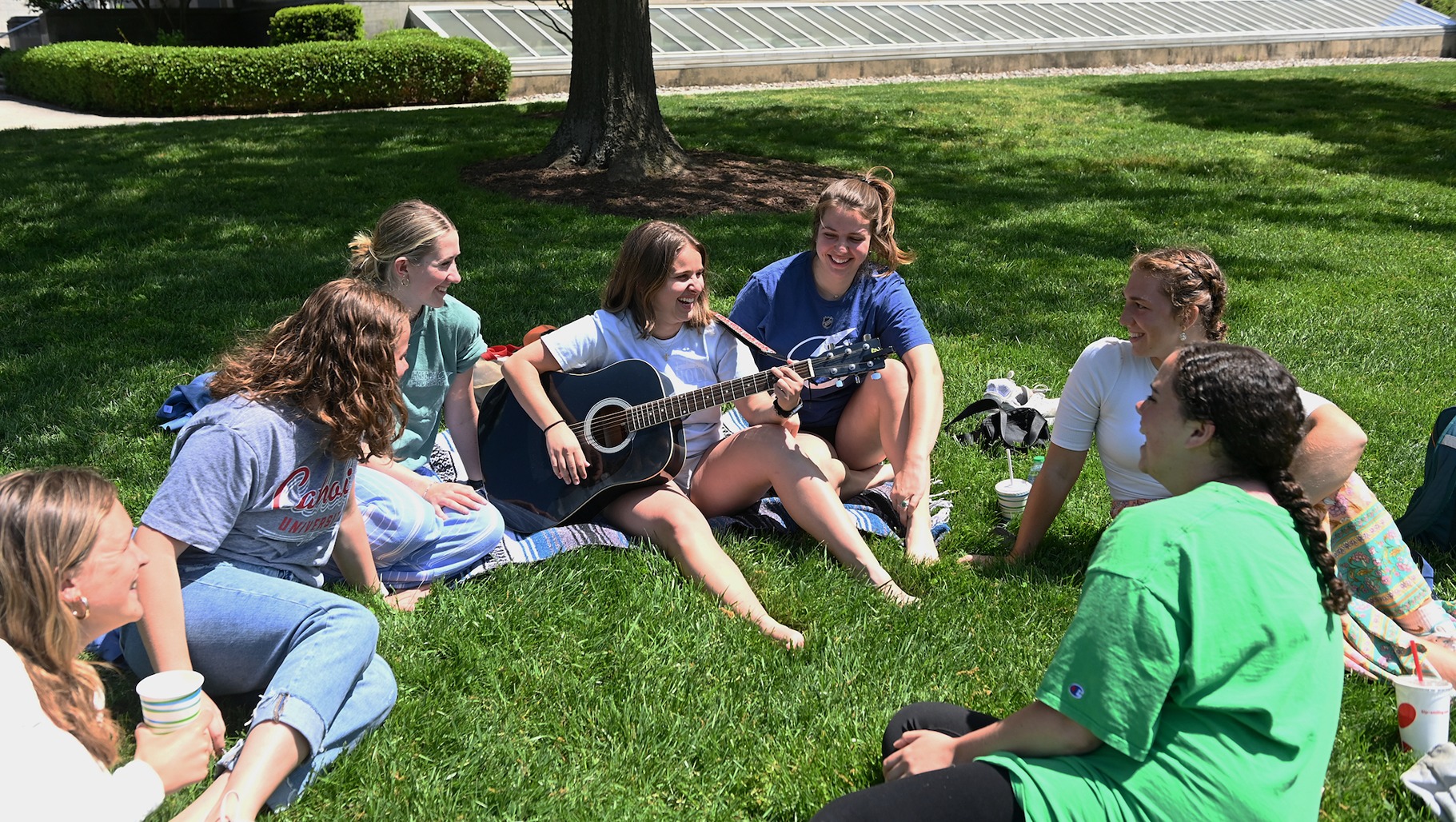 student plays the guitar on a lawn on campus with group of friends gathered around her