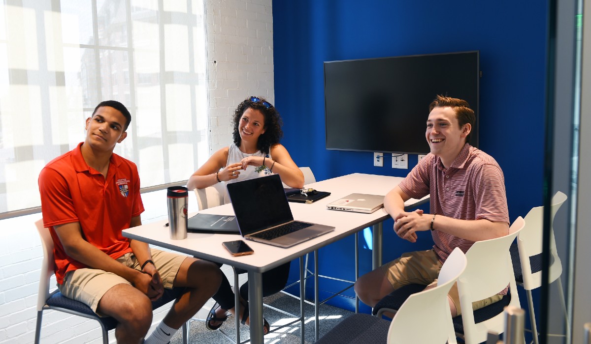 Three students are smiling at something off camera. They are sitting in one of the Busch School's study bubbles.