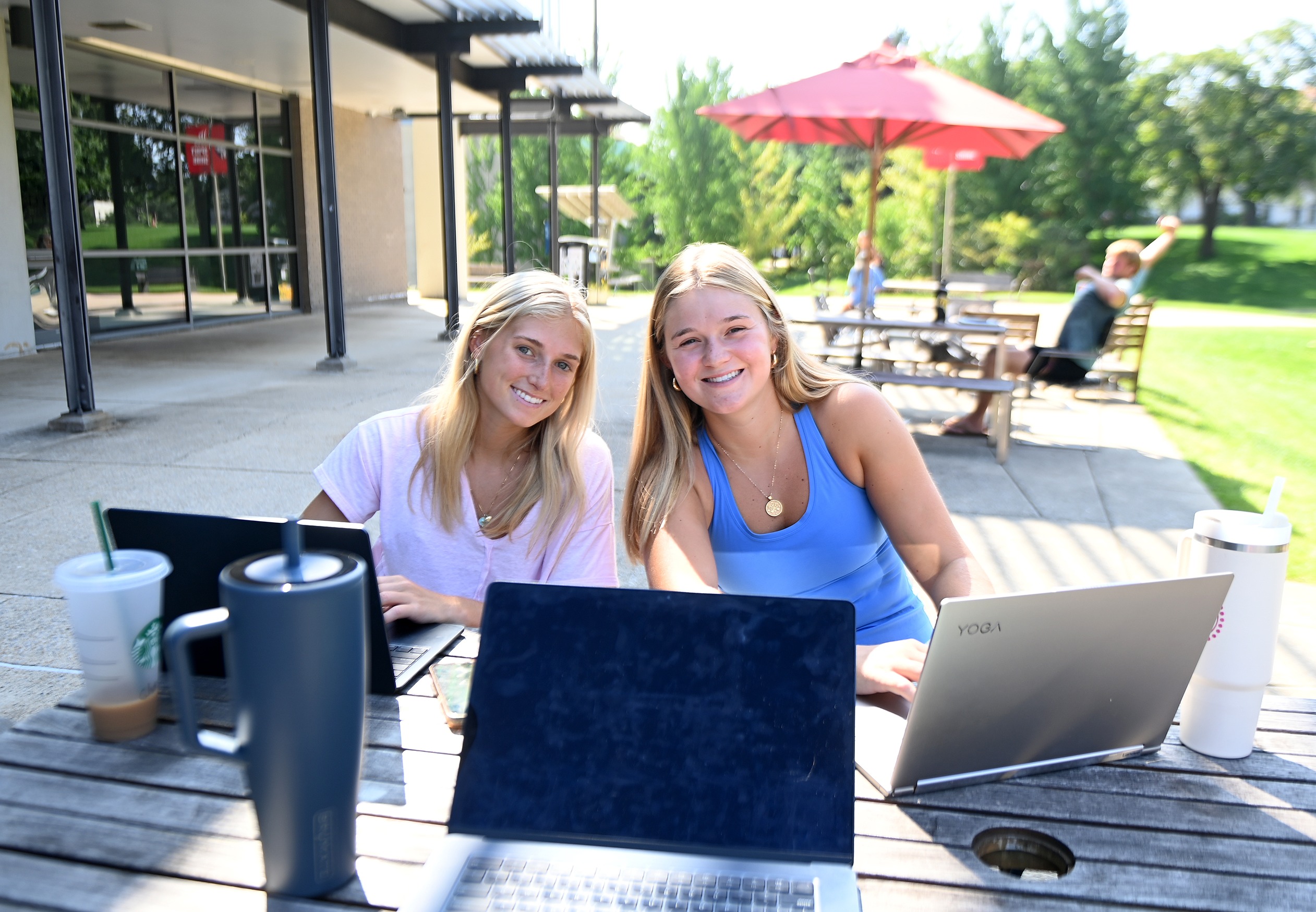 two girls smile at the camera as they work on their laptops outdoors