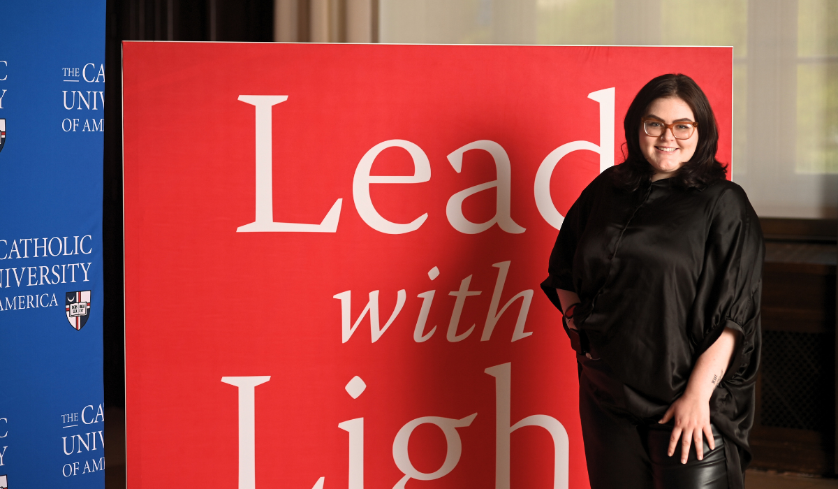 Olivia is standing infront of a red Lead With Light background. She is wearing a black shirt and pants.